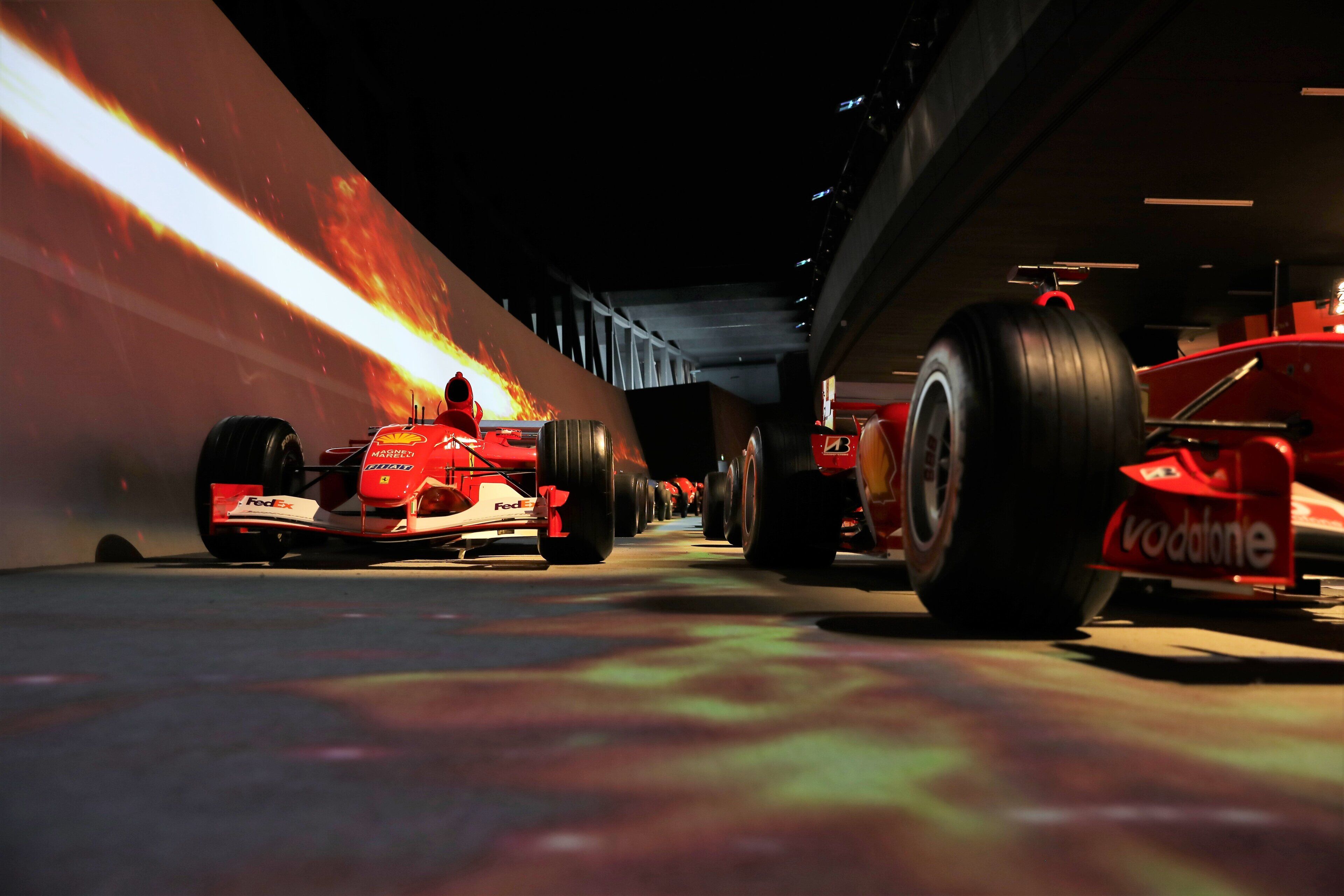A row of red Formula 1 cars parked indoors with a large screen displaying a fiery animation in the background.