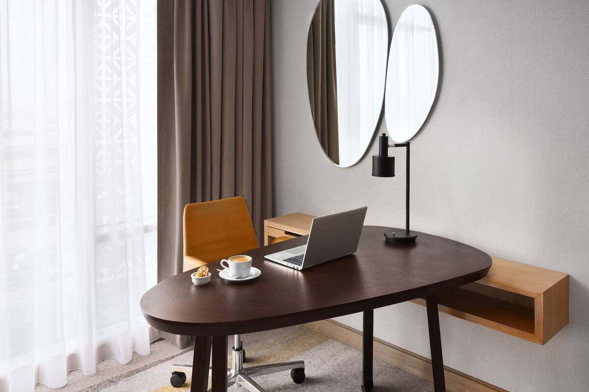 Une chambre d’hôtel moderne avec une table ronde, un ordinateur portable, une tasse de café et une petite assiette de biscuits. On y trouve également une chaise, un miroir et une étagère en bois.