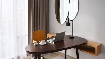 Habitación de hotel moderna con una mesa redonda, una computadora portátil, una taza de café y un plato pequeño de galletas. Hay una silla, un espejo y un estante de madera.