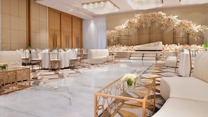 Un hall de hotel espacioso y elegantemente decorado, con una zona de estar central, varias mesas y un arreglo floral decorativo.