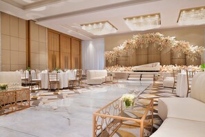 Un hall de hotel espacioso y elegantemente decorado, con una zona de estar central, varias mesas y un arreglo floral decorativo.
