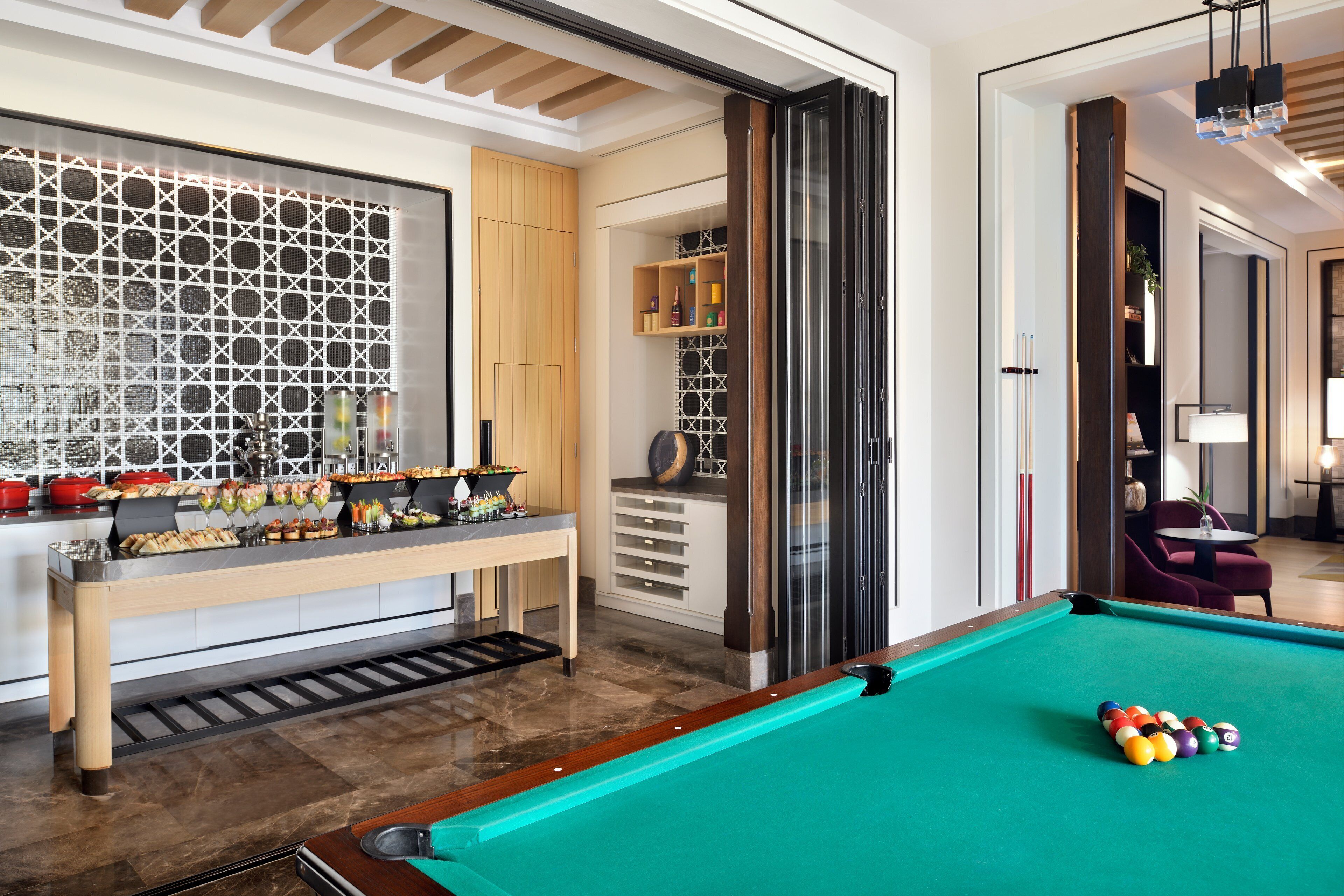 Une table de billard recouverte d’un feutre vert, accompagnée d’un jeu de billes. On y trouve également un banc en bois avec un plateau en dessous, et un mur orné d’un motif géométrique.