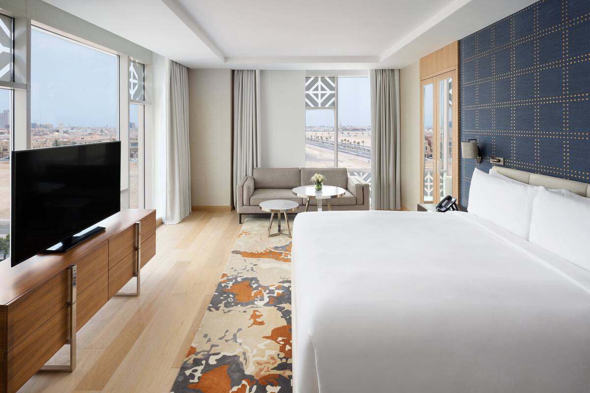 Une chambre d’hôtel avec un grand lit, un téléviseur posé sur une console en bois, un canapé, une petite table ronde et une vue sur la ville grâce à de grandes fenêtres.