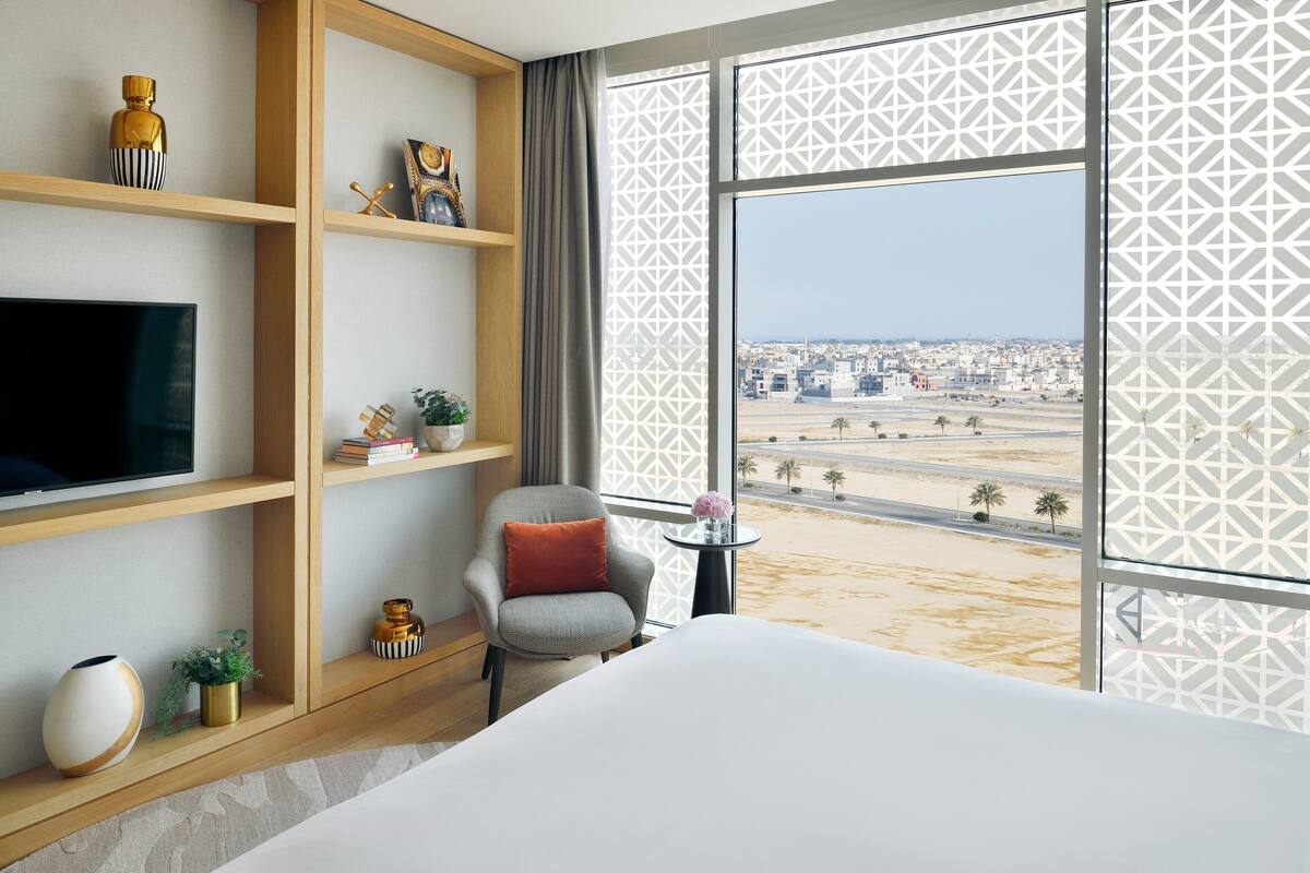 Quarto de hotel com uma cama grande, televisão, uma poltrona com almofada, mesa pequena com um vaso e vista para a paisagem urbana através de cortinas estampadas.