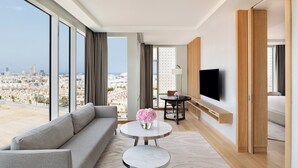 A modern hotel room with a large window offering a city view, a grey sofa, a round table with a vase of flowers, a flat-screen TV mounted on the wall, and a wooden wardrobe.