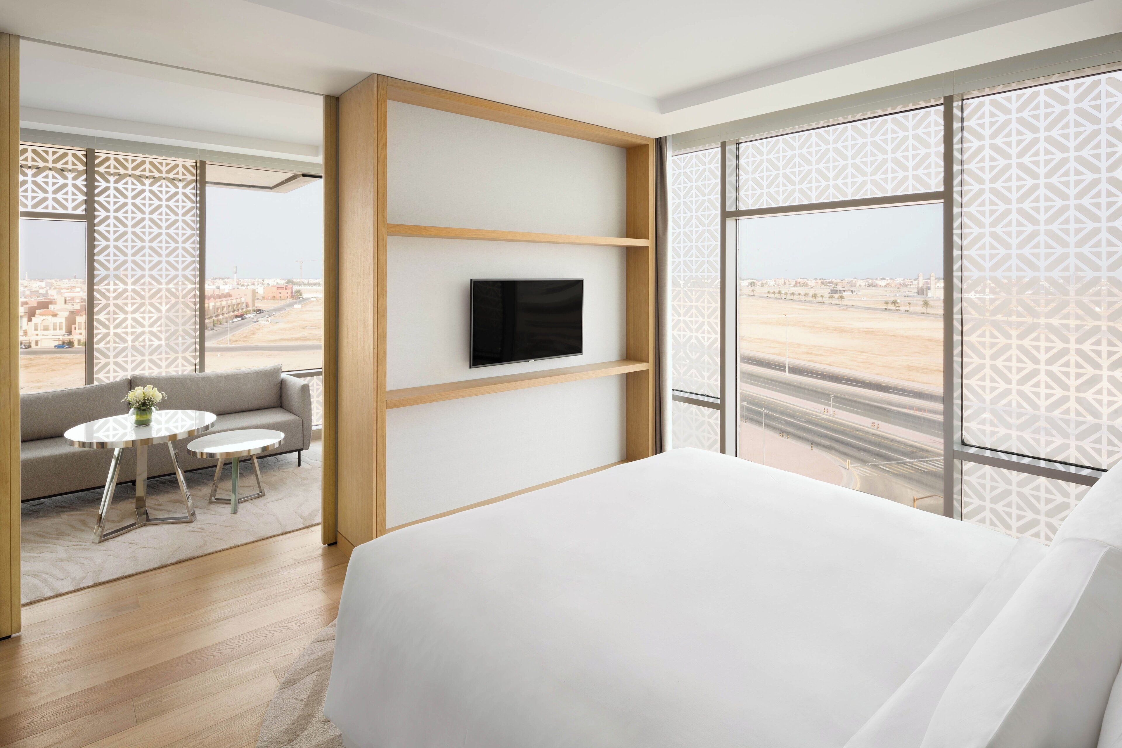 Een moderne hotelkamer met een groot bed, een televisie bevestigd aan een houten plank, een bank, twee kleine tafels en een uitzicht op een stadsgezicht door grote ramen.