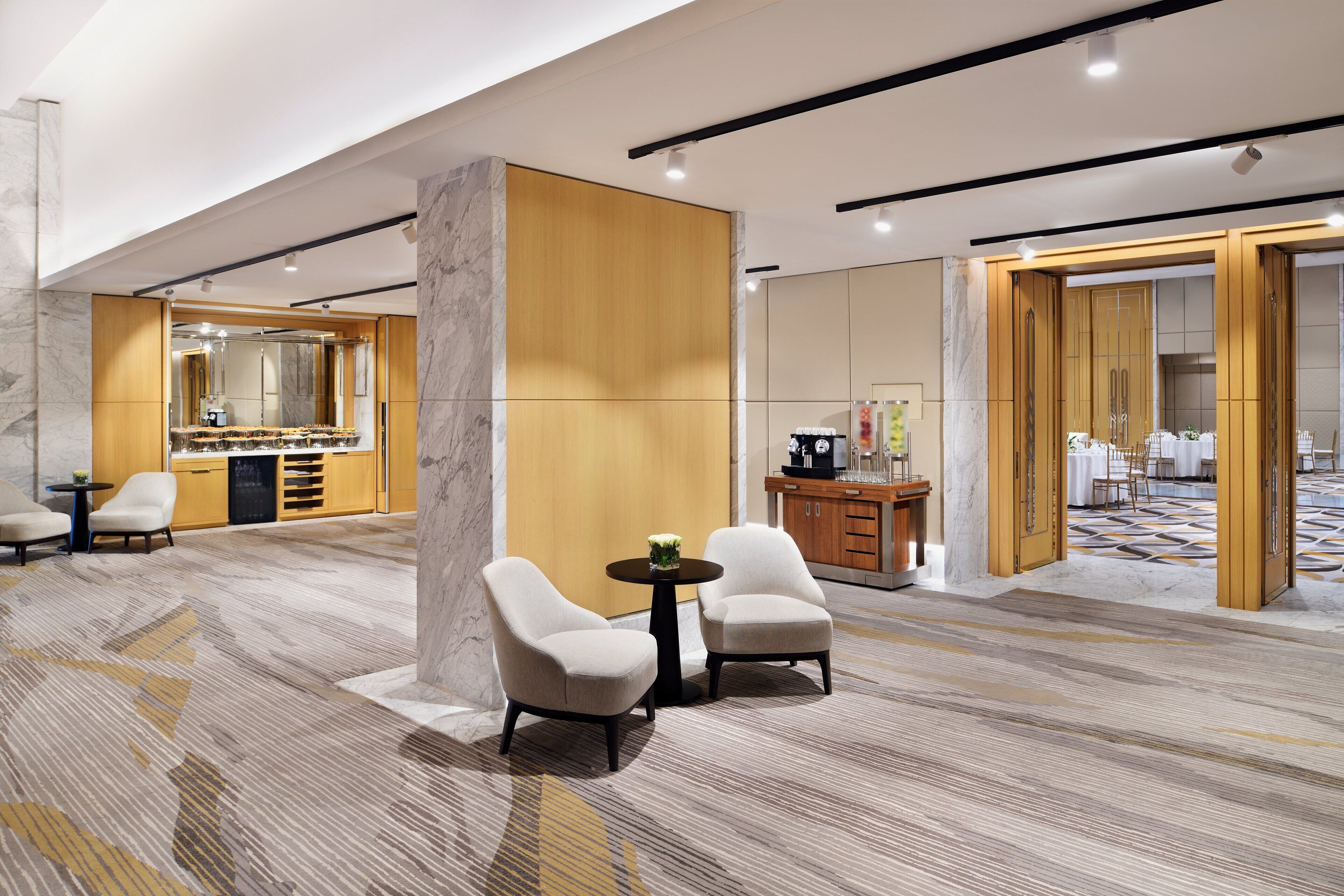 A modern hotel lobby with a marble floor, two armchairs, a small round table, a wooden cabinet, and a large mirror.