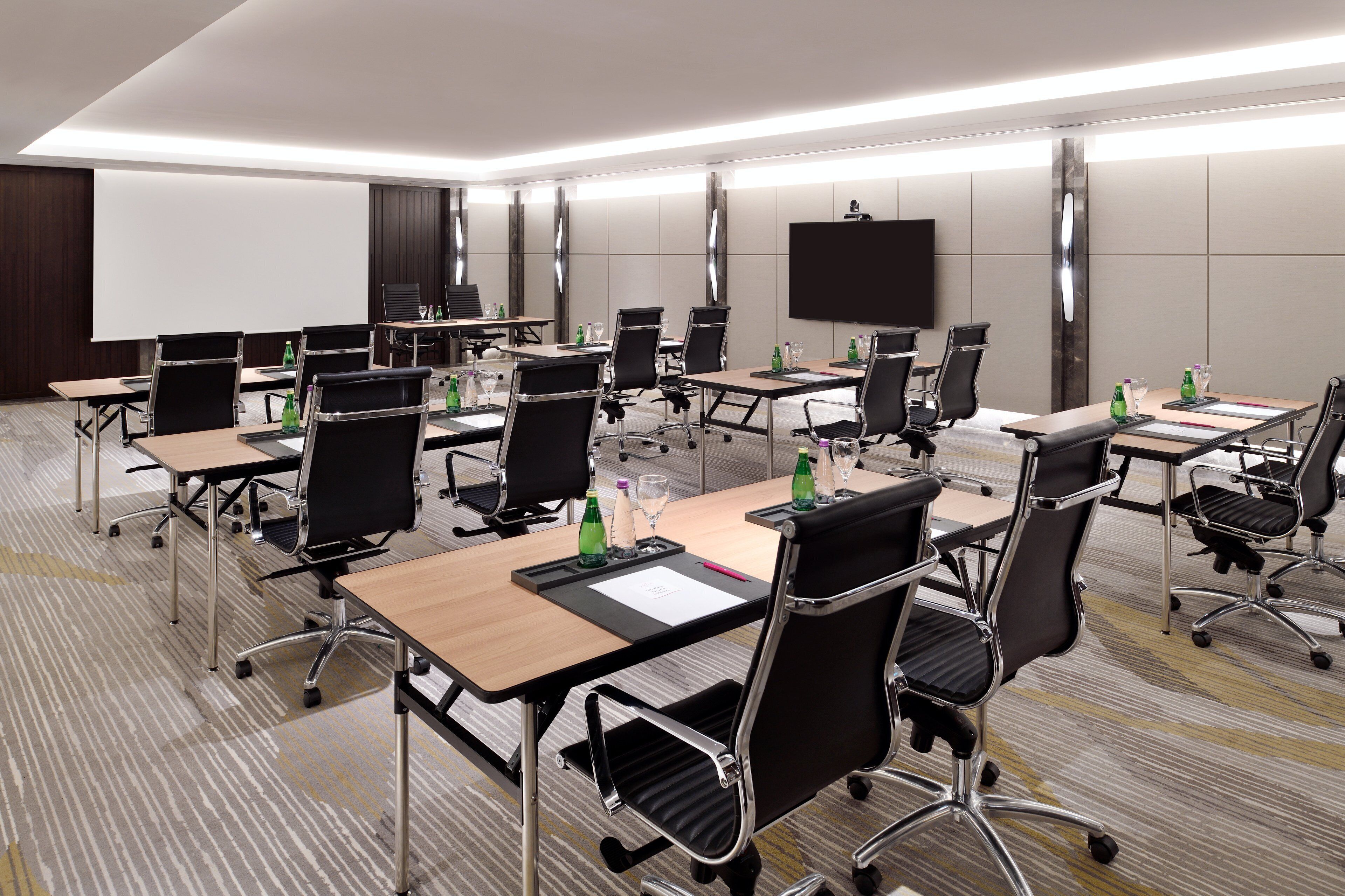 A modern conference room with multiple tables and chairs, equipped with water bottles and glasses, and a large screen on the wall.
