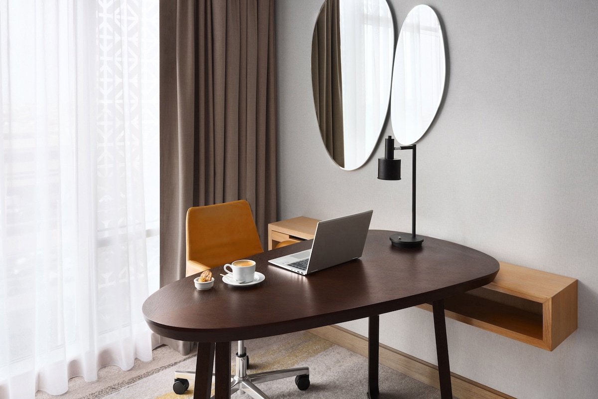 A modern hotel room with a round desk, a laptop, a cup of coffee, and two mirrors on the wall.
