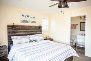 A bedroom with a bed, a nightstand, a ceiling fan, a bathroom with a toilet and sink, and a motivational poster on the wall. - Evergreen Lane Getaway · River Views and Front Porch Sitting in Paradise (Livingston)