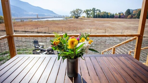 A wooden table with a vase of yellow flowers on a balcony overlooking a field and mountains. - Evergreen Lane Getaway · River Views and Front Porch Sitting in Paradise (Livingston)
