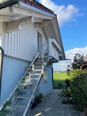 A white building with a metal staircase leading to a door, surrounded by greenery and a clear sky.