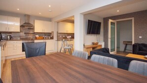 Een moderne keuken met witte kasten, een eethoek met een houten tafel en stoelen, en een zithoek met een bank en televisie.