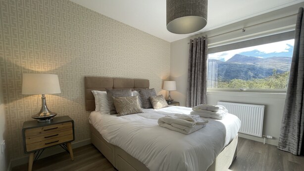 Een moderne slaapkamer met een groot bed, twee nachtkastjes met lampen, en uitzicht op bergen door het raam, met behangpapier met een patroon.