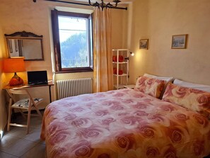 A bedroom with a large bed, a desk with a laptop, a chair, a radiator, a window with curtains, and a shelf with towels. - Apartment in an old barn, private garden overlooking the village (Plesio)