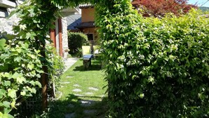 Une maison en pierre avec un chemin de jardin menant à une porte, entourée d’une végétation luxuriante et avec une colline en arrière-plan.