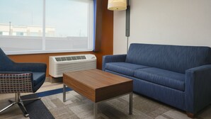 A modern hotel room with a blue sofa, a wooden coffee table, a patterned armchair, and a large window with blinds.
