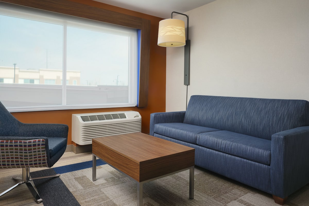A modern hotel room with a blue sofa, a wooden coffee table, a patterned armchair, and a large window with blinds.