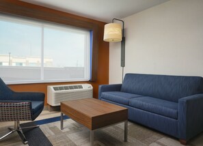 Et moderne hotelværelse med en blå sofa, et træbord, en mønstret lænestol og et stort vindue med persienner.