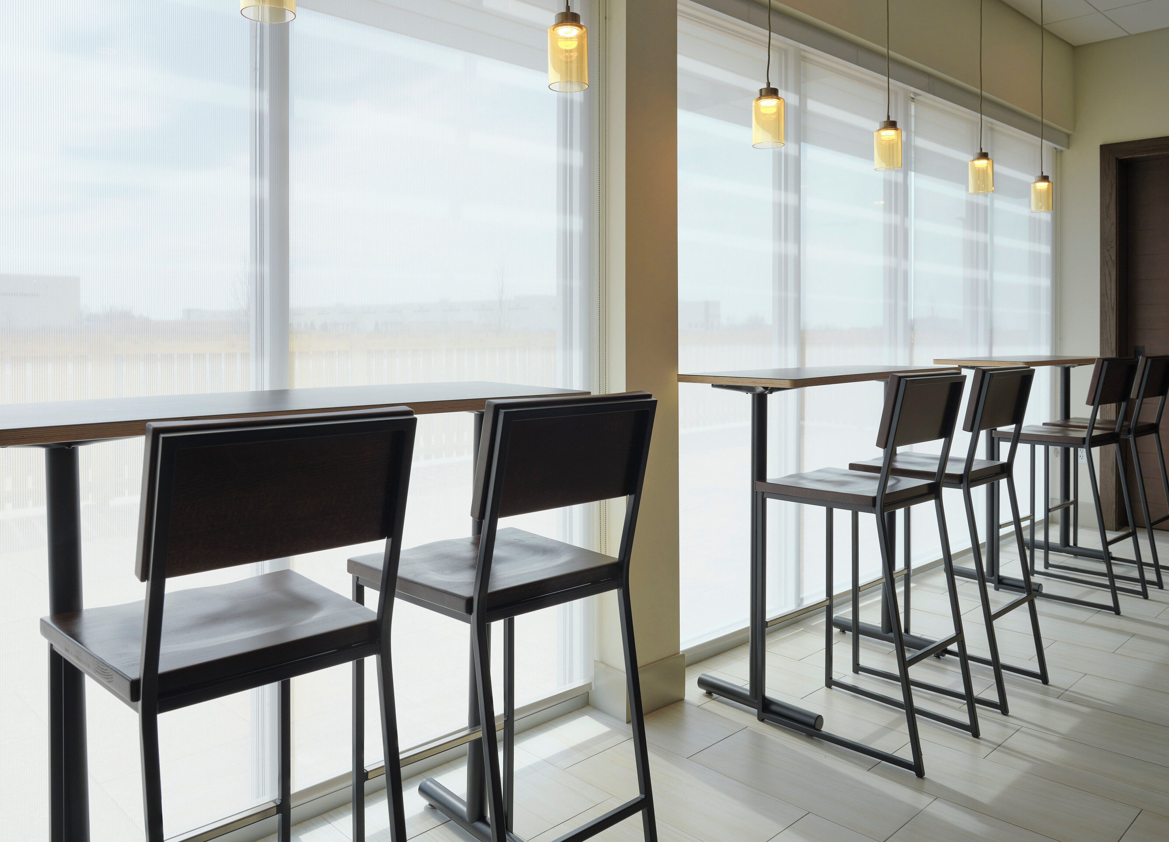 A modern interior with a row of tables and chairs, large windows with blinds, and pendant lighting.