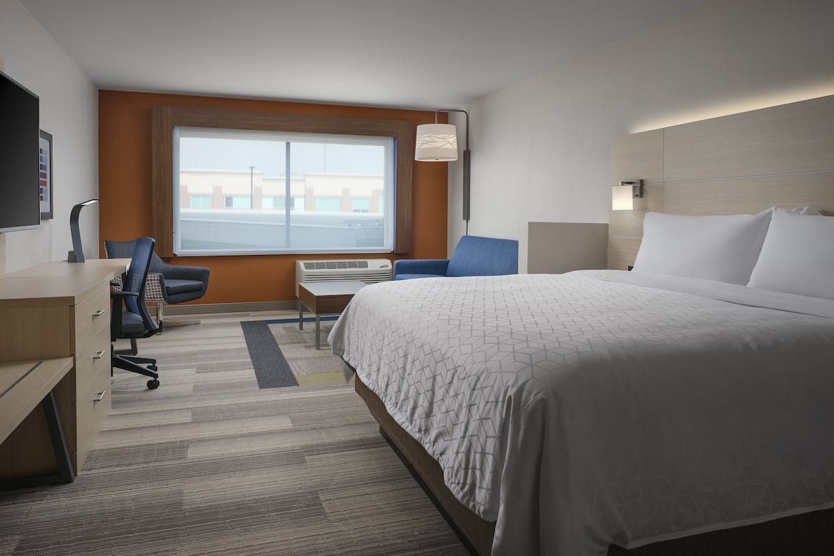A hotel room with a large bed, a desk with a chair, a window with a view of buildings, and a wall-mounted lamp.