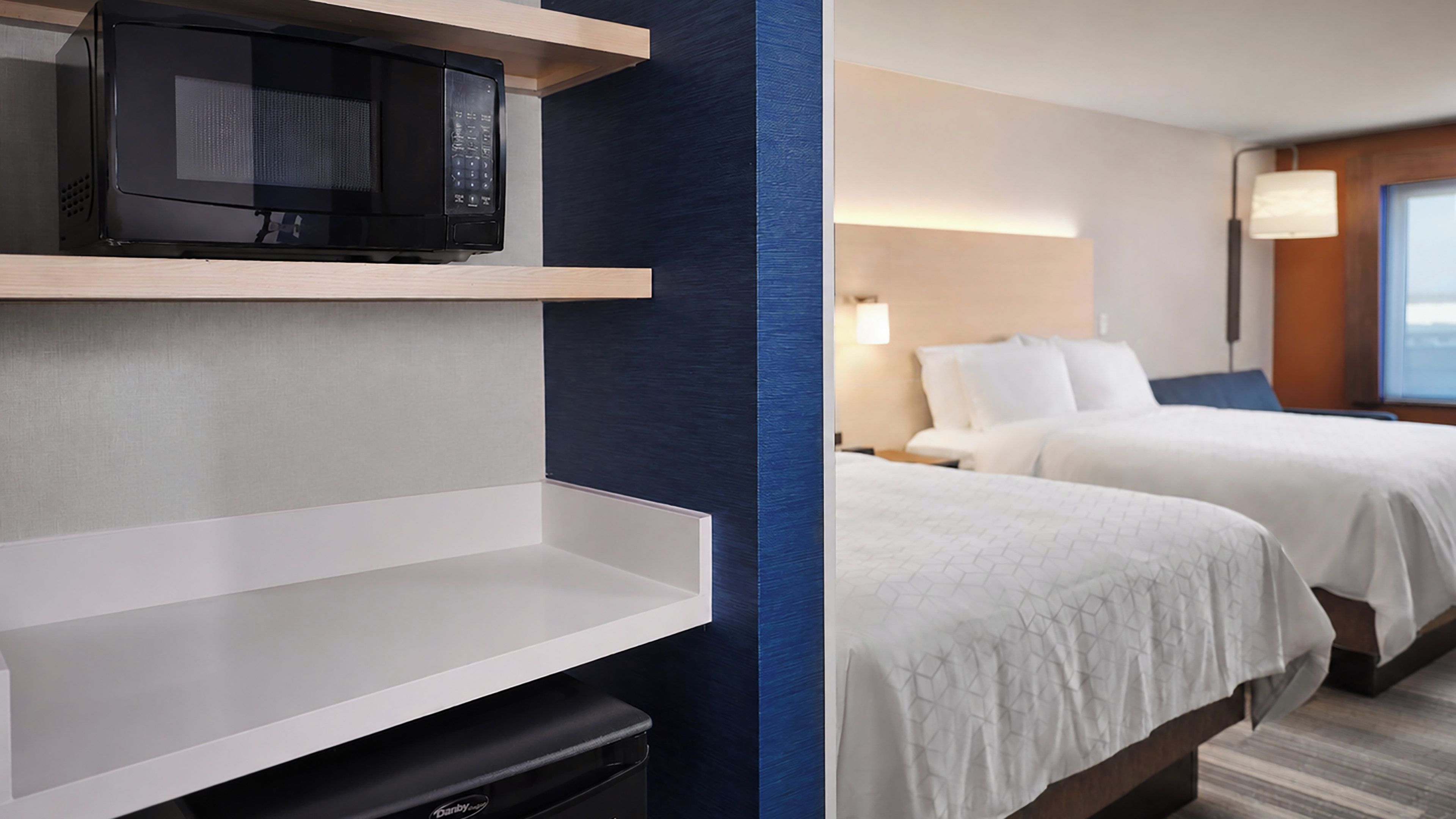 A hotel room with a microwave on a shelf, a bed with white bedding, and a nightstand with a lamp.