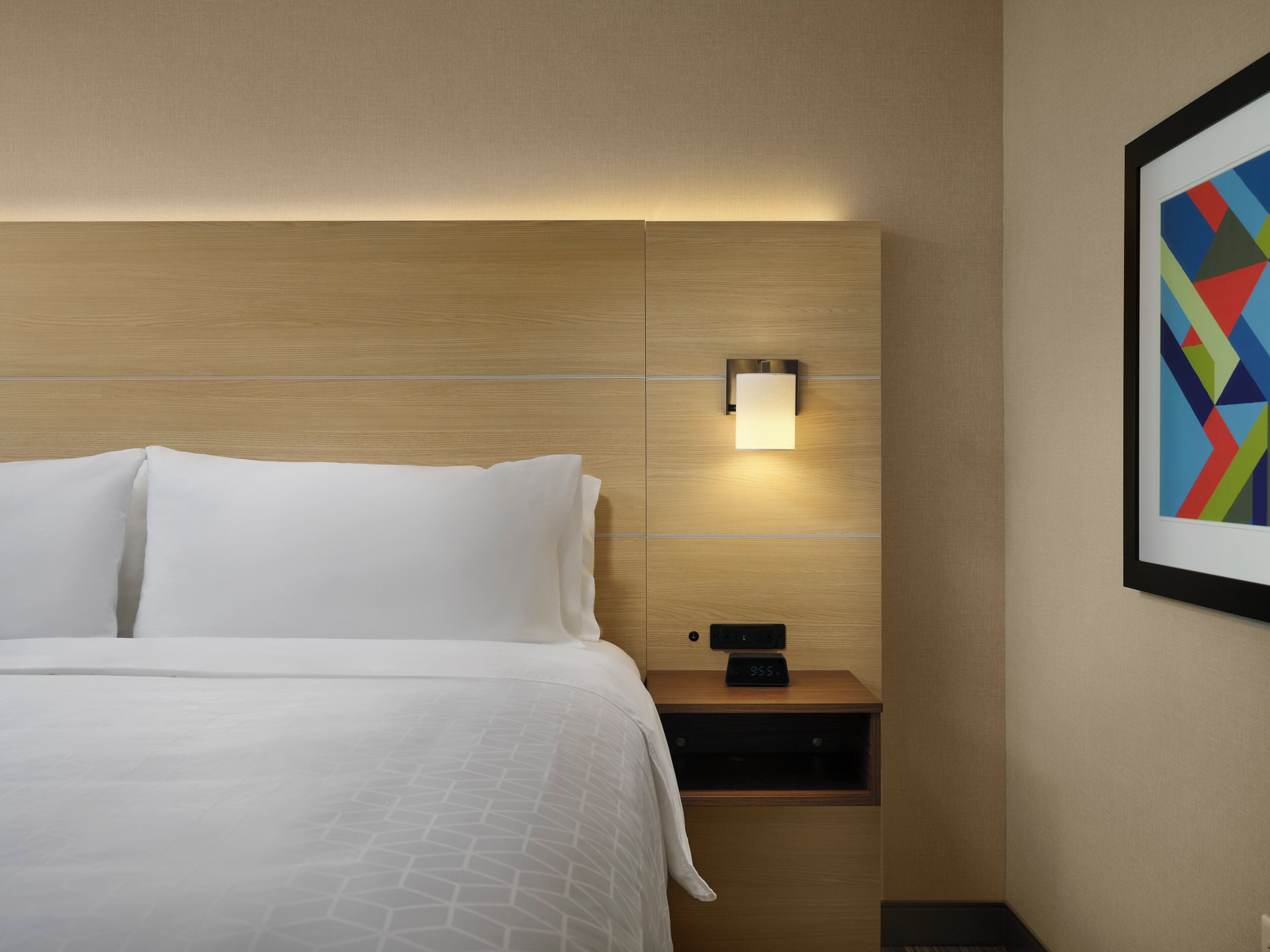 A hotel room with a bed, a nightstand with a clock, and a wall-mounted light fixture.
