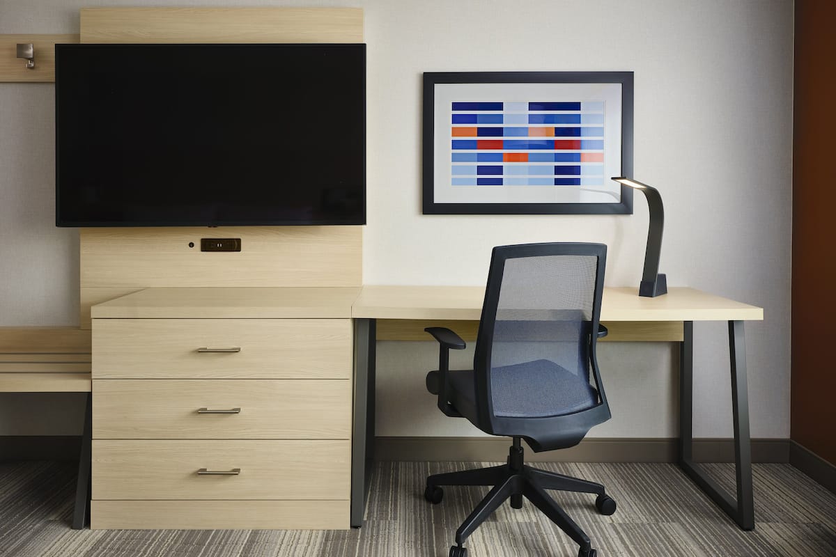 A modern office desk with a flat-screen monitor, a framed abstract artwork, a black office chair, and a desk lamp.