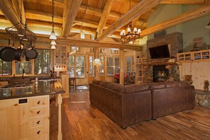A rustic cabin interior with a stone fireplace, wooden beams, a leather sofa, and a kitchen with hanging pots.