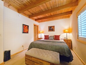 Een slaapkamer met een houten plafond, een bed met een groene deken, twee rode kussens, twee nachtkastjes met lampen, en een raam met jaloezieën.