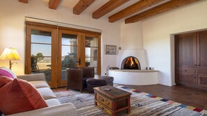 Una sala de estar acogedora con chimenea, vigas de madera, un sofá, sillones y vistas a través de puertas de cristal.