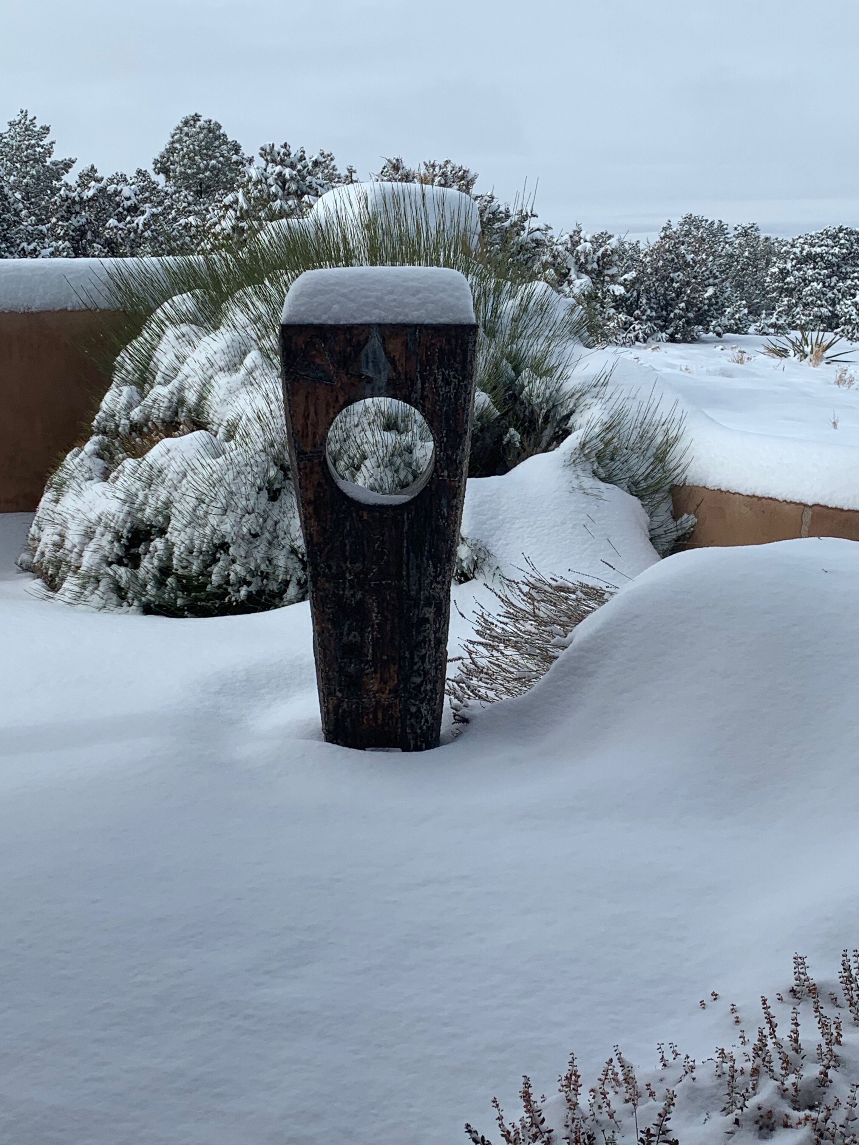 A snow-covered planter with a circular cutout, surrounded by snow and sparse vegetation.