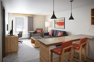A modern hotel room with a wooden desk, a sofa with cushions, a dining table with chairs, and pendant lighting.
