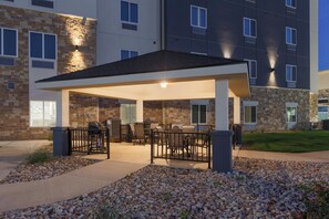 A covered outdoor seating area with tables and chairs, a stone building with multiple windows, and a well-maintained gravel pathway. - Candlewood Suites San Antonio - Schertz (Schertz)