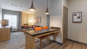 A modern hotel room with a kitchenette, a dining area, a sofa, and a TV.