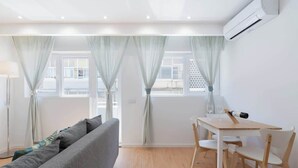 Ein moderner, gut beleuchteter Raum mit einem grauen Sofa, einem Holzespeisetisch mit Stühlen und großen Fenstern mit leichten Vorhängen.