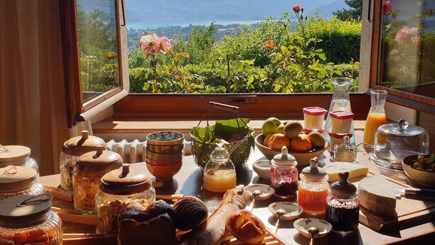 Une table de petit-déjeuner avec divers aliments et condiments, une fenêtre donnant sur un paysage verdoyant et des montagnes.