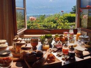 Une table de petit-déjeuner avec divers aliments et condiments, une fenêtre donnant sur un paysage verdoyant et des montagnes.