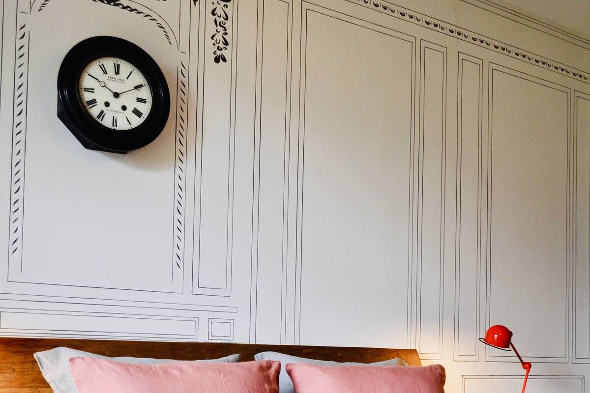 Un lit bien fait, avec des oreillers roses et blancs, une horloge au mur, une lampe rouge sur une table de chevet, et un plateau avec un verre de jus d’orange et une bouilloire.