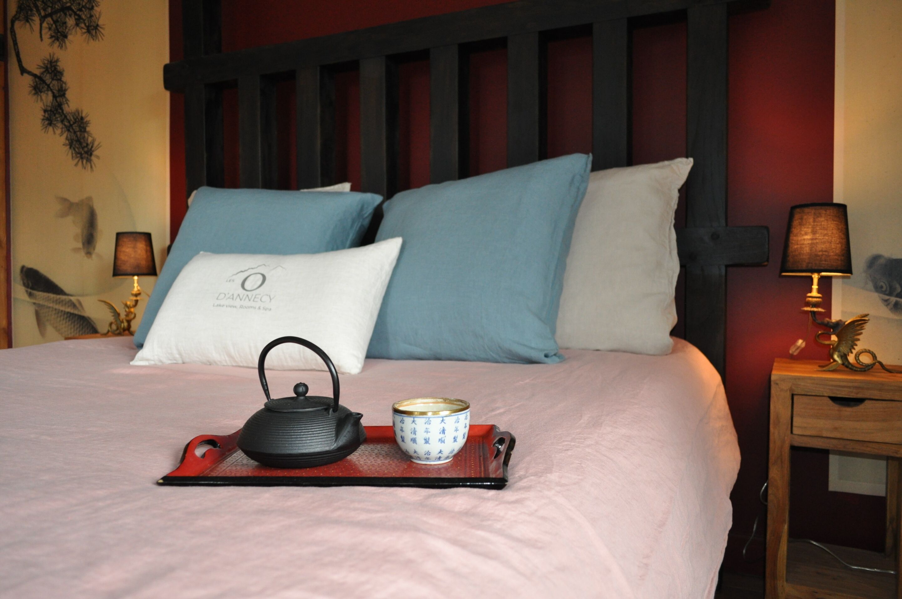 Un lit bien fait, avec un plateau contenant du thé, des coussins, et un mur agrémenté d’un élément décoratif.