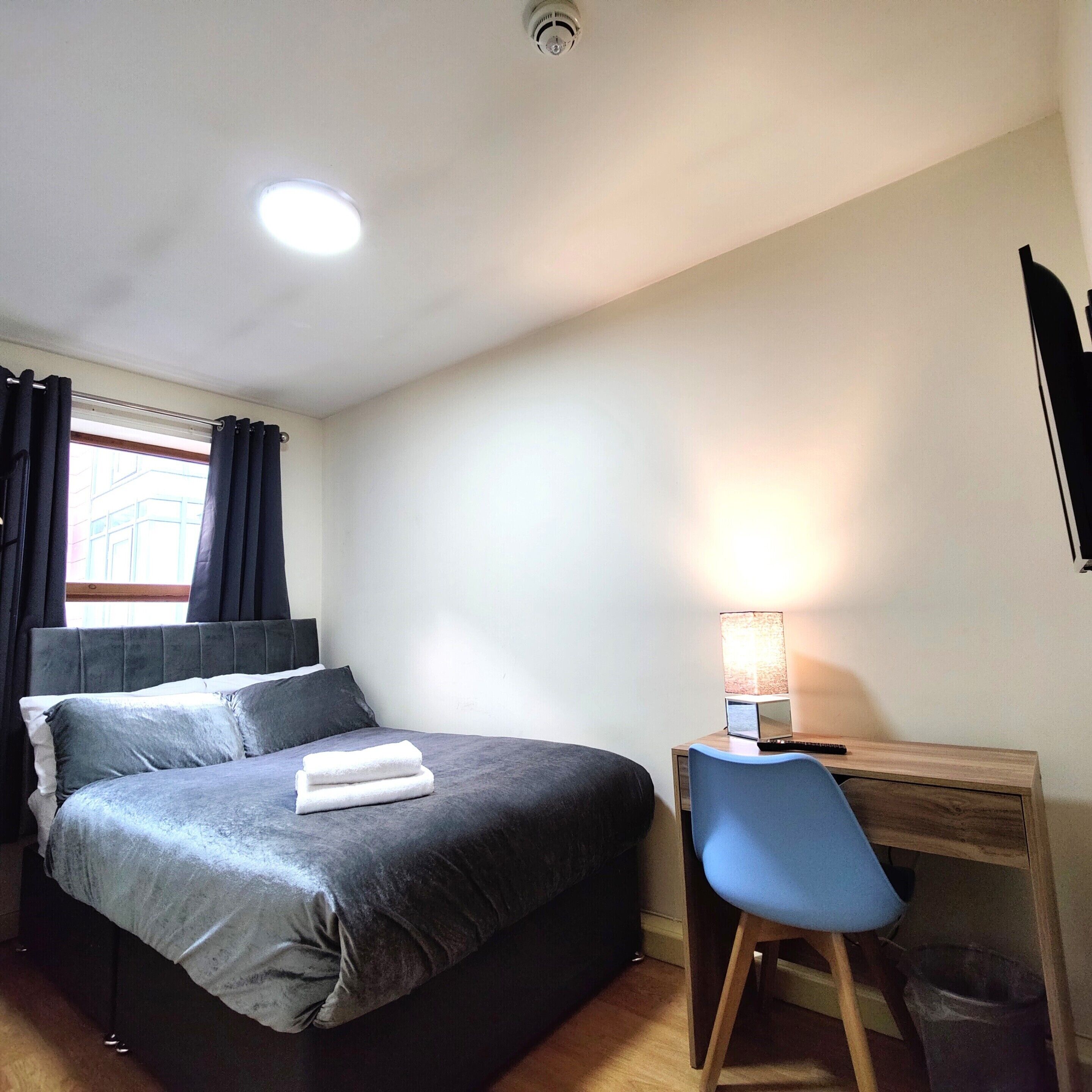 A bedroom with a bed, a desk, a chair, a lamp, and a window with curtains.