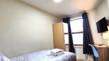 A small, neatly arranged room with a bed, a desk with a chair, a television, and a window with curtains.