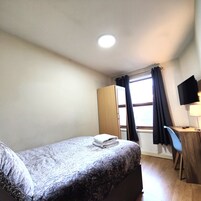 A small, neatly arranged room with a bed, a desk with a chair, a television, and a window with curtains.