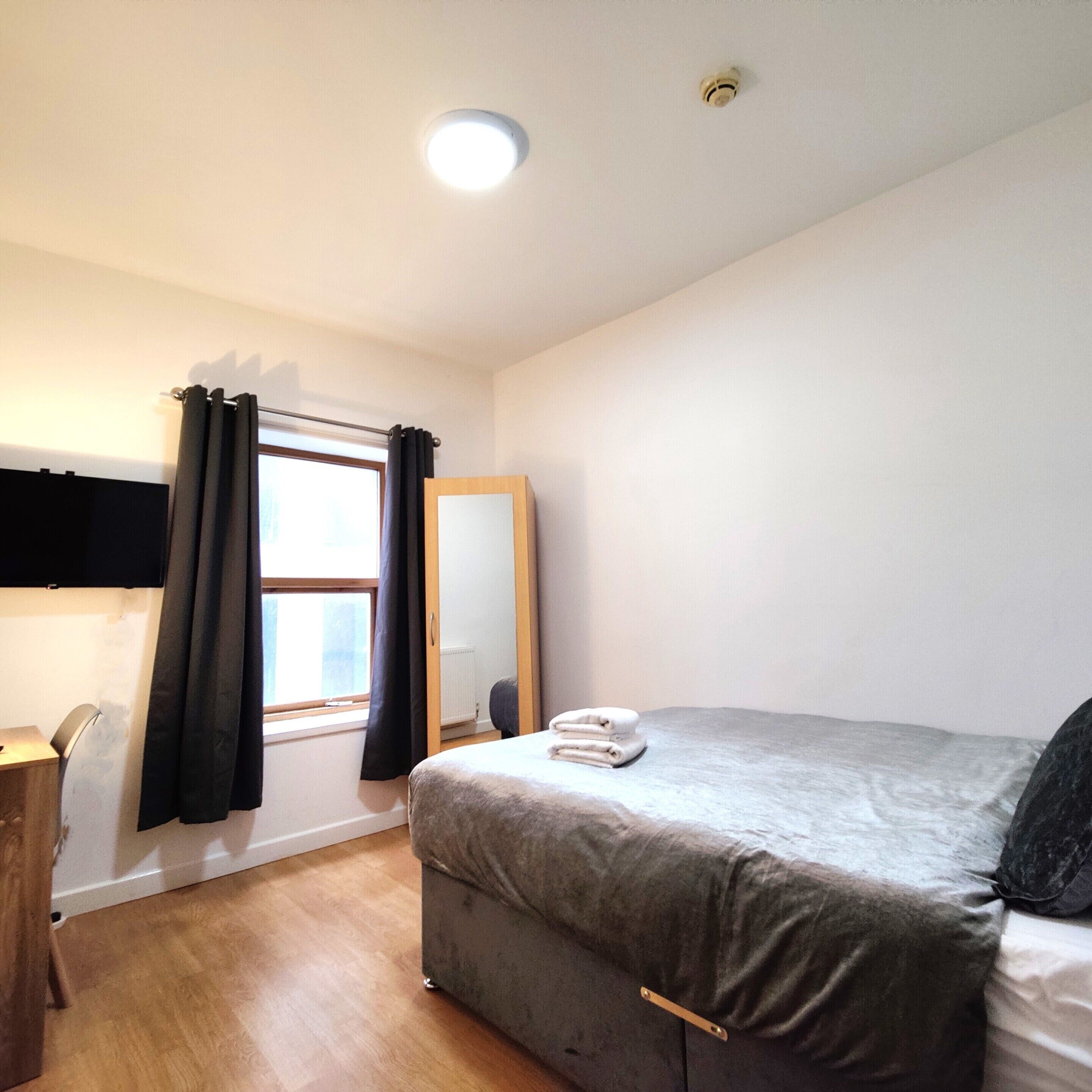 A bedroom with a bed, a bedside table, a window with curtains, a mirror, and a ceiling light.