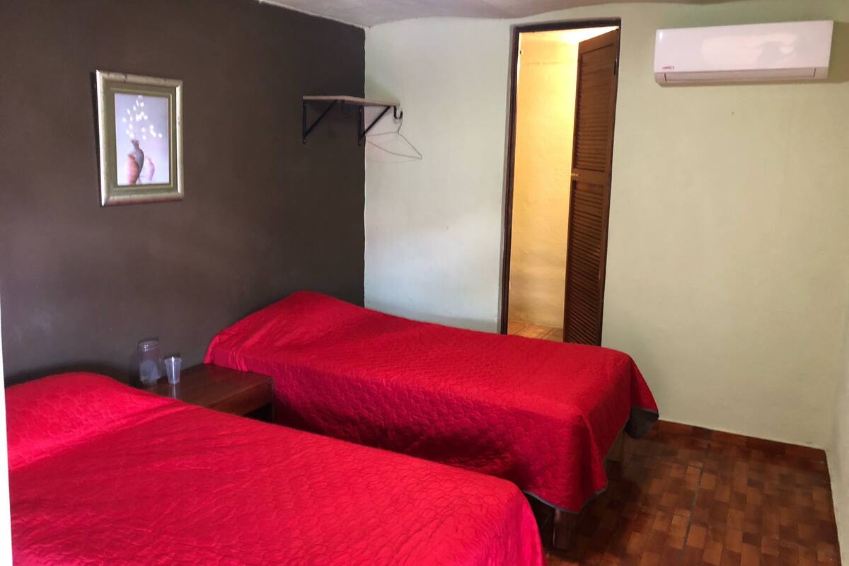 Een kamer met twee bedden, een klein tafeltje, een wandairconditioner en een deur naar een andere kamer.