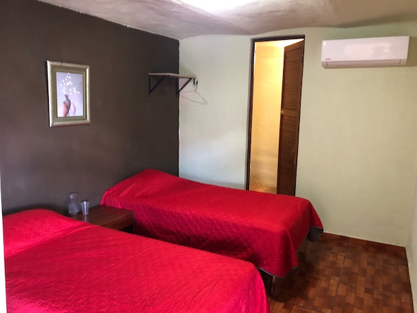 A room with two beds, a small table, a wall-mounted air conditioner, and a door leading to another room.