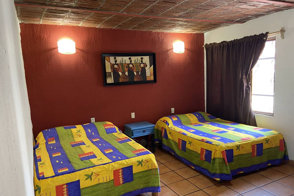 Een kamer met twee bedden, elk bedekt met kleurrijke quilts, een schilderij aan de muur en een raam met gordijnen.