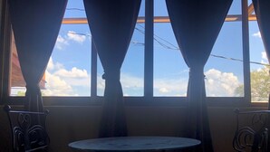 A room with a round table and two chairs, a window with curtains, and a view of the sky and clouds.