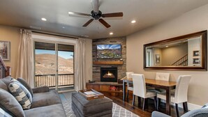 A living room with a fireplace, a dining area, and a balcony with a view of a mountainous landscape.