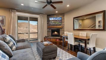 A living room with a fireplace, a dining area, and a balcony with a view of a mountainous landscape.